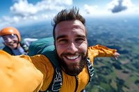 Adventurous bearded man in yellow jacket taking selfie while skydiving with instructor against scenic mountain landscape and blue cloudy sky background.