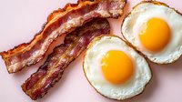 Crispy fried bacon strips and sunny side up eggs with bright yellow yolks on pink background, classic breakfast combination photographed from above.