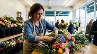 Young woman with long dark hair and fair complexion, wearing denim shirt and apron, arranging colorful flowers at wooden counter in busy flower shop with customers and blooming plants in background.