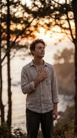 Caucasian male in casual linen shirt standing under tree canopy at golden hour, gazing upward with hand on chest in contemplative pose by water.