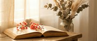 Open book with pink cherry blossoms on a rustic wooden table, dried eucalyptus in a glass vase, warm sunlight through sheer curtains.