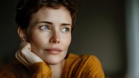 Close-up portrait of contemplative woman with short curly hair in mustard sweater against dark background, hand resting on chin in thoughtful pose.