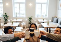 Three diverse young professionals toasting with coffee cups in a bright, modern office break room with wooden tables, potted plants, and natural light from large windows.