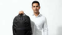 Young man with dark hair and olive complexion wearing white long-sleeve shirt holding black laptop backpack with multiple compartments. Isolated on white background for design use.