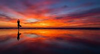 Silhouette of person practicing yoga on wet beach at sunset with dramatic orange and purple sky reflecting in water creating mirror effect for wellness content.