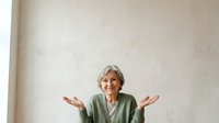 Mature woman with gray hair wearing olive green cardigan gestures with open palms in welcoming or uncertain pose against neutral beige wall background.