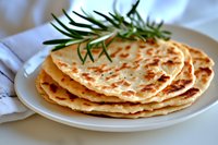 Stack of homemade golden flatbread with crispy spots and fresh rosemary sprig garnish on white plate against light background, close up food photography.