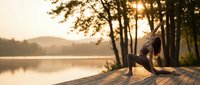 Young woman practicing yoga warrior pose on wooden dock at sunrise by peaceful lake surrounded by trees creating serene morning wellness atmosphere.