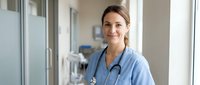 Smiling female healthcare professional in blue scrubs with stethoscope standing in modern hospital corridor with medical equipment in background.