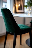 Elegant emerald green velvet dining chair with black legs against blurred modern interior background featuring soft bokeh lights and minimalist decor elements.