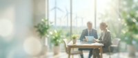 Blurred business meeting with two professionals discussing at wooden table in bright modern office with large windows and green plants creating bokeh effect.