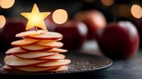Creative holiday dessert made from sliced red apples arranged in Christmas tree shape topped with glowing star candle against dark background with bokeh lights.