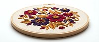 Embroidered floral design on cream fabric stretched in wooden hoop, featuring burgundy, purple, and gold thread flowers arranged in circular pattern for needlework and craft projects.