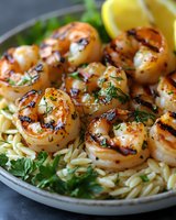 Grilled shrimp with charred marks served over orzo pasta, garnished with fresh herbs and lemon wedges on gray plate, close up view of seafood dinner.