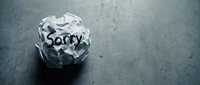Crumpled paper ball with the word Sorry written on it, lying on a dark gray surface, symbolizing regret, apology, and failed communication.