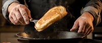 Senior male baker with weathered hands tossing freshly baked bread loaf over hot cast iron skillet, flour dust flying, artisan baking process and traditional bread making technique.