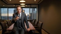 Confident middle-aged Caucasian businessman in gray suit pointing forward in modern boardroom with city skyline view during golden hour meeting.