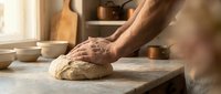 Baker kneading fresh bread dough on floured wooden surface in warm artisan kitchen with natural lighting and traditional baking tools.