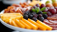 Appetizing charcuterie board with sliced cheddar cheese, prosciutto, salami, fresh blackberries and red grapes arranged on white ceramic plate, macro food photography.