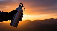 Hand holding stainless steel water bottle against dramatic mountain sunset sky with orange and purple clouds for outdoor adventure and hydration concept.