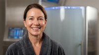 Middle-aged Caucasian woman smiling warmly in modern kitchen wearing gray robe, natural morning light creating cozy domestic atmosphere for lifestyle content.