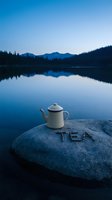 Vintage enamel teapot on a lakeside rock with TEA inscription at dusk, calm blue water and mountain silhouettes in background.