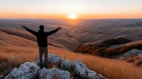 Male hiker with raised arms celebrating sunrise over golden rolling hills landscape. Freedom, achievement, outdoor adventure concept for motivational content.