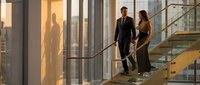 Professional business couple in formal attire walking down modern glass staircase in corporate office building during golden hour sunset lighting.