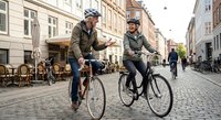 Senior couple cycling together on cobblestone street in historic city center, wearing helmets and enjoying active retirement lifestyle outdoors.