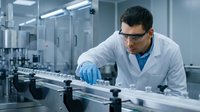Male scientist in white lab coat and safety glasses examining samples in modern laboratory setting with equipment and machinery.