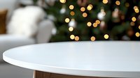 Minimalist white round table against blurred Christmas tree background with soft golden bokeh lights creating cozy festive atmosphere in modern interior.