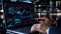 Financial analyst pointing at trading charts and market data on laptop screen in dark office with blue data visualization and stock market graphs.