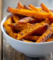 Roasted sweet potato wedges seasoned with herbs and spices served in white ceramic bowl on rustic wooden table, close-up view of healthy vegetable side dish.