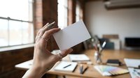 Hand holding blank white business card in modern office workspace with natural lighting for branding and corporate identity mockup design projects.