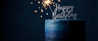 Elegant dark blue birthday cake with a sparkling sparkler and silver Happy Birthday topper, glowing against a deep navy background.
