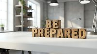 BE PREPARED wooden letters on desk in modern office workspace with shelving and natural light, symbolizing readiness and business planning.