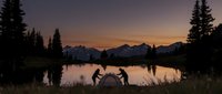 Two hikers setting up tent at mountain lake during golden hour sunset with snow-capped peaks and evergreen forest silhouettes reflecting in calm water.