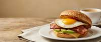 Gourmet breakfast burger with fried egg, crispy bacon and fresh lettuce on sesame bun served on white plate with coffee cup on wooden table background.