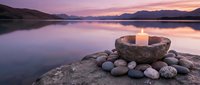 Stone bowl with burning candle surrounded by smooth pebbles on lakeside at sunset creating peaceful meditation and spa atmosphere for wellness relaxation.