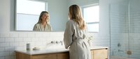 Young woman in bathrobe looking at mirror reflection in modern bathroom with white subway tiles and natural light for morning skincare routine.