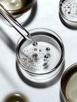 Clear liquid droplets falling from dropper into glass petri dish creating spherical bubbles against white background, macro photography with dramatic shadows.