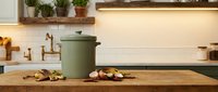 Modern kitchen interior with green ceramic pot on wooden countertop, brass faucet, white subway tiles, floating shelf with plants and warm lighting.