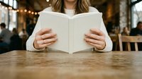 Woman with long brown hair reading open book at wooden table in café with warm lighting and blurred background, suitable for education and leisure concepts.