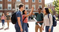 Diverse college students celebrating success with high five on university campus courtyard during sunny day with historic brick buildings in background.