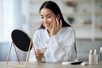 Young Asian woman applying skincare cream while looking in mirror during morning beauty routine at home vanity table with cosmetic products.
