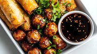 Asian appetizer platter with glazed meatballs, spring rolls, sesame seeds and soy dipping sauce garnished with fresh green onions on white ceramic plate.