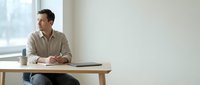 Young professional man sitting at wooden desk in modern minimalist office space looking thoughtfully out window during daytime work break.