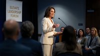 Professional businesswoman in white suit speaking at corporate leadership conference with engaged audience listening to presentation.