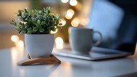 Small green plant in white ceramic pot on wooden stand next to coffee cup and laptop with soft bokeh lights creating cozy home office atmosphere.
