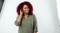 Happy African American woman with curly burgundy hair talking on phone in olive green shirt against white studio background for business content.